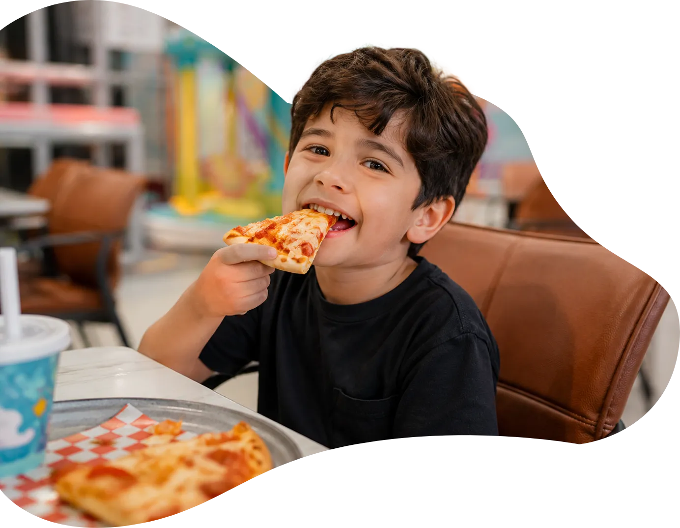 Playtropolis-kid-eating-pizza-cafe-woodbridgewebp - playtropolis Smiling young boy in a black shirt taking a bite of pizza at a table in a casual restaurant.