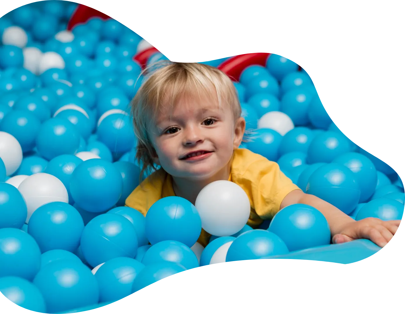 Playtropolis-toddler-ball-pit-woodbridgewebp - playtropolis Smiling toddler in a yellow shirt lies in a ball pit filled with blue balls, reaching toward the camera.